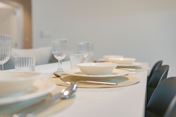 Una mesa de comedor dispuesta con platos, fuentes y cubiertos blancos, con un mantel individual y otro tejido debajo.