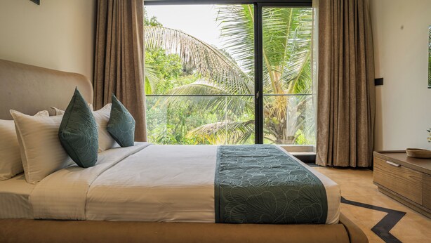A bedroom with a large bed, a view of palm trees through a sliding glass door, and a wooden dresser. - Lovely 3-bedroom villa in Saligao with WiFi access (Saligao)