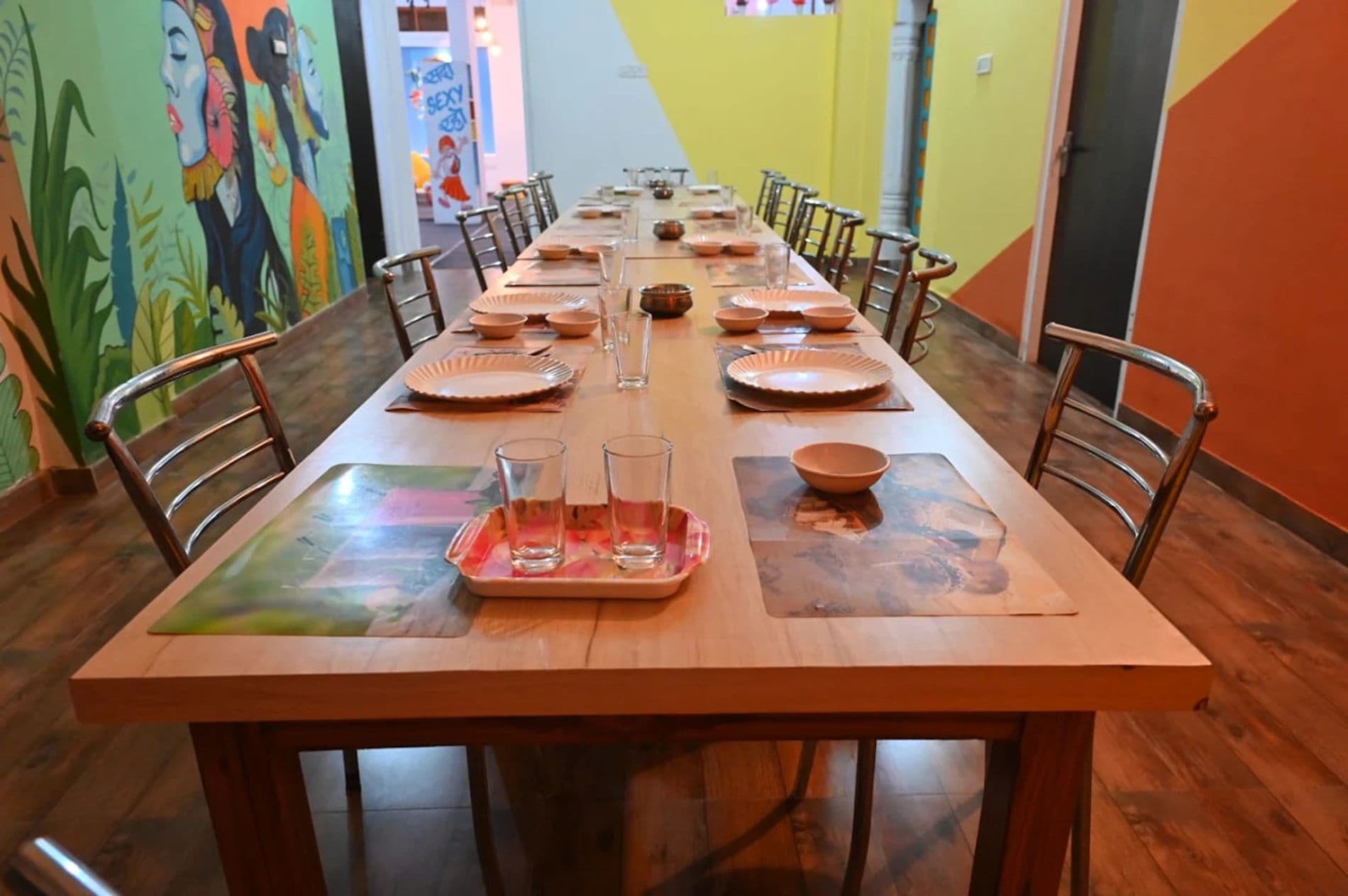 Una larga mesa de comedor preparada para varias personas con sillas a ambos lados, en una habitación con paredes coloridas y una puerta visible al fondo.