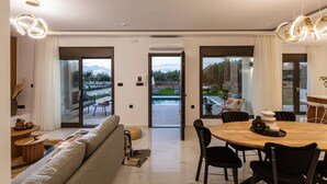 Ein modernes Wohnzimmer mit einer Couch, einem Esstisch und Stühlen. Große Fenster und eine Klimaanlage sind an der Wand zu sehen.