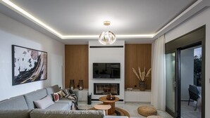 Ein modernes Wohnzimmer mit grauem Sofa, einem Flachbildfernseher, einem Kamin und einem großen Fenster mit Blick ins Freie.