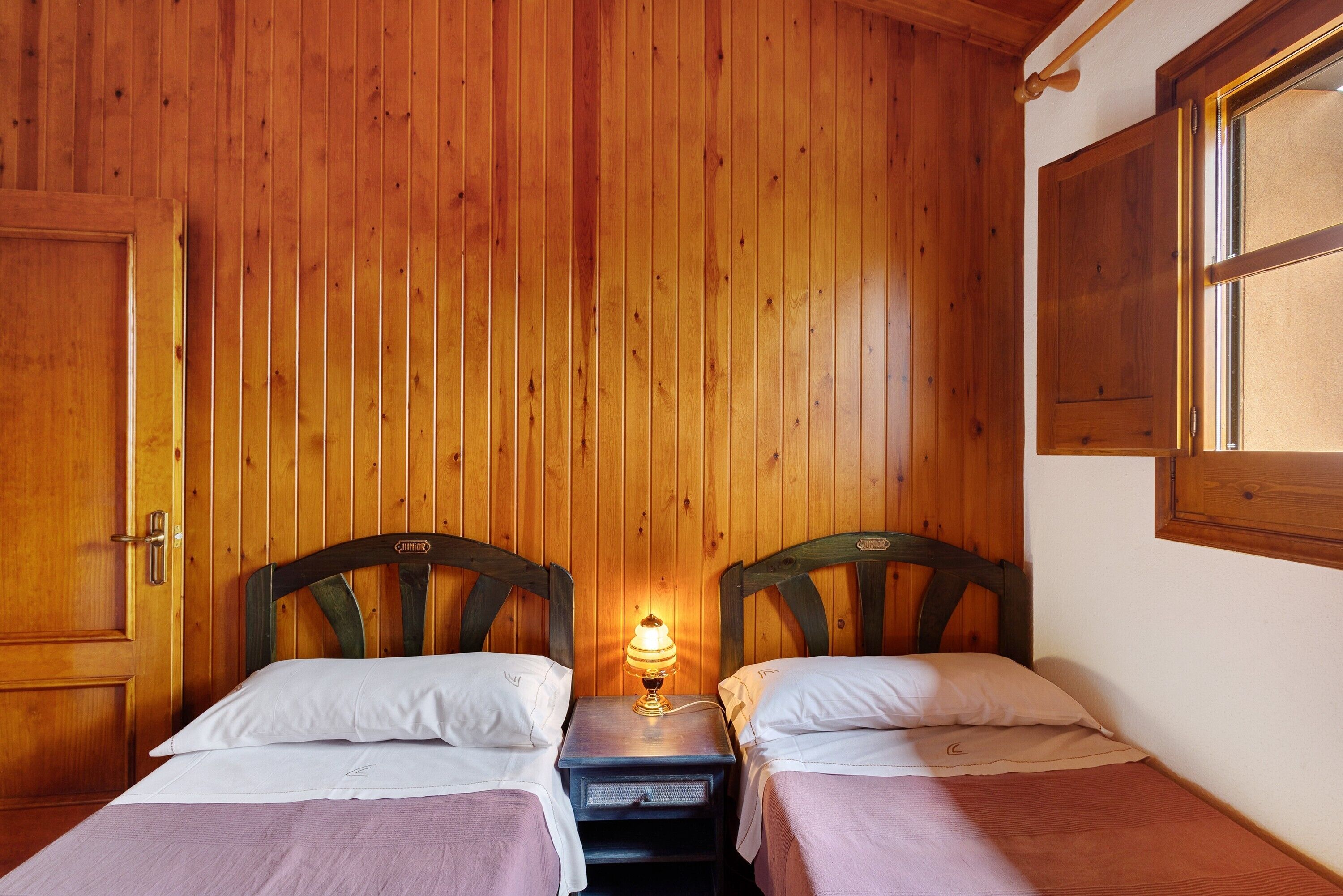 Ein Zimmer mit zwei Holzbettgestellen, einem Nachttisch mit Lampe, Holzwänden und einem Fenster.