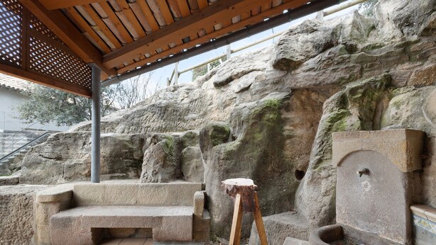 Un banc en pierre, un tabouret en bois et une fontaine en pierre, le tout installé dans un espace extérieur avec un mur rocheux en arrière-plan.