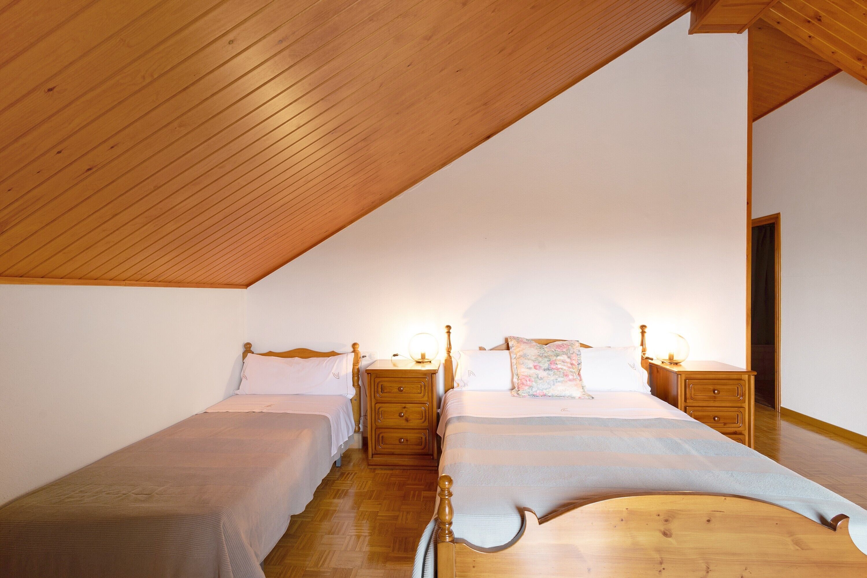 Un dormitorio con dos camas individuales, cabeceros de madera, mesitas de noche con lámparas y un techo inclinado.