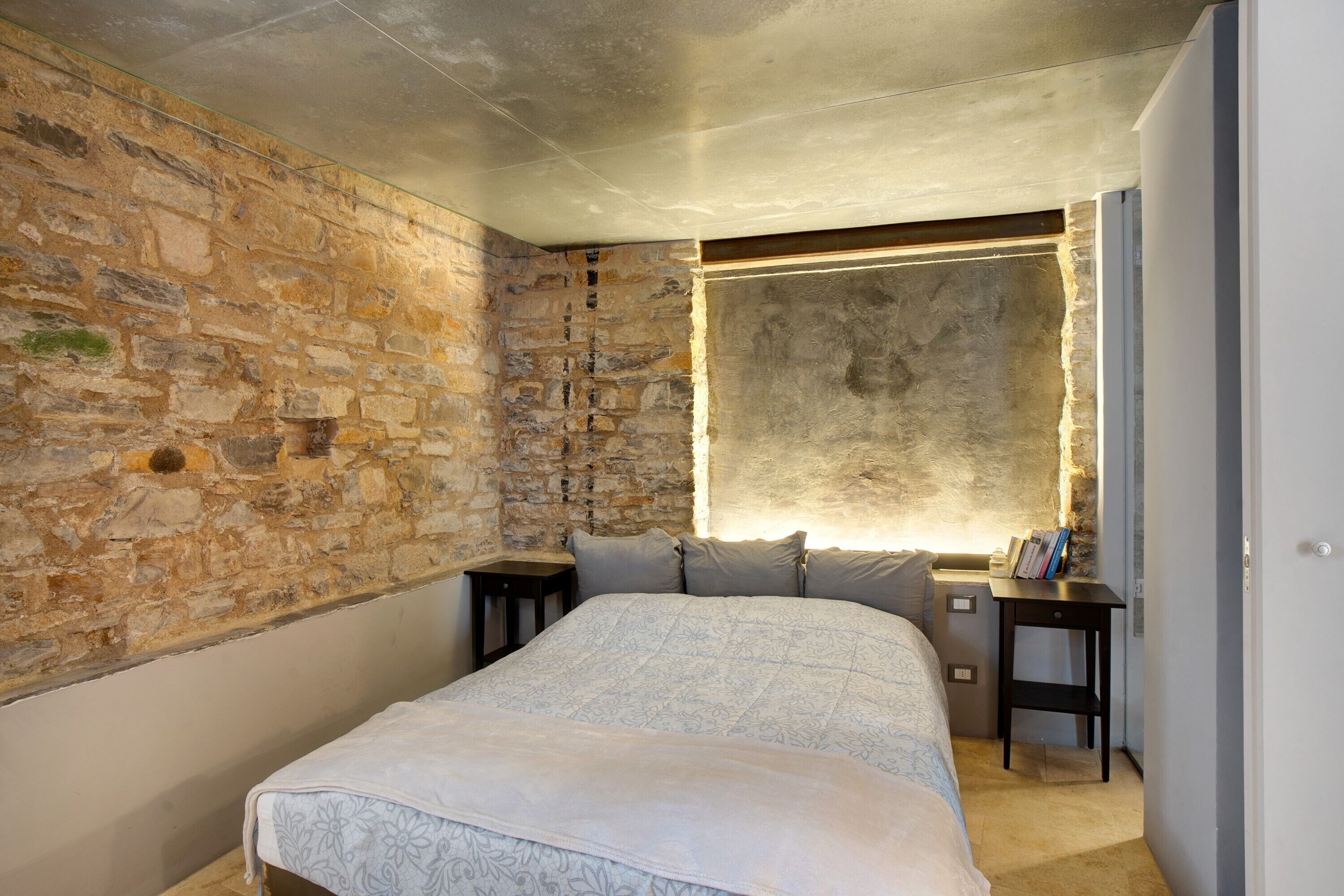 Quarto com parede de pedra, cama com colcha estampada, mesa de cabeceira com livros e criado-mudo.