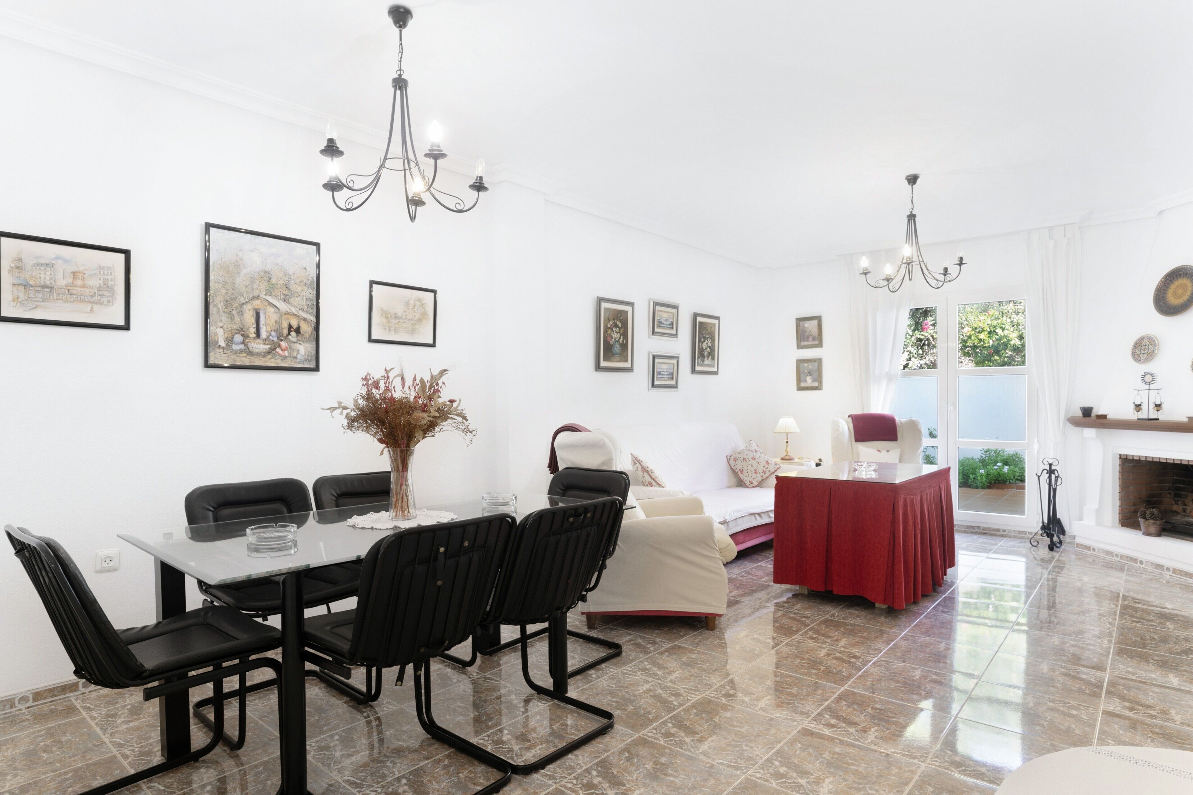 A dining area with a table, chairs, and a chandelier. There are framed pictures on the walls and a fireplace in the background.