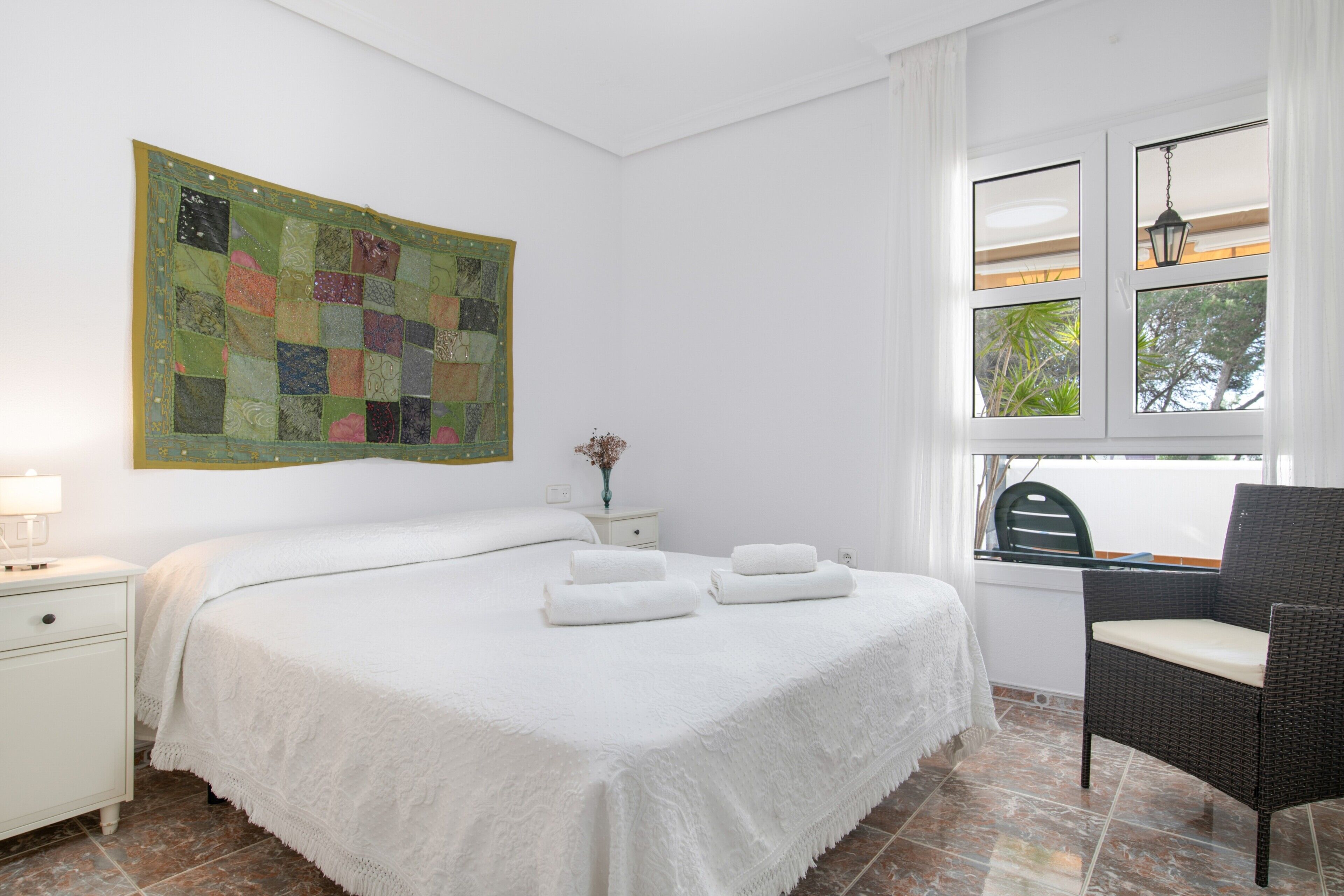 A neatly arranged bedroom with a large bed, a chair, a nightstand, a wall hanging, and a window with a view.