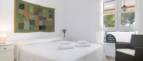 A neatly arranged bedroom with a large bed, a chair, a nightstand, a wall hanging, and a window with a view.