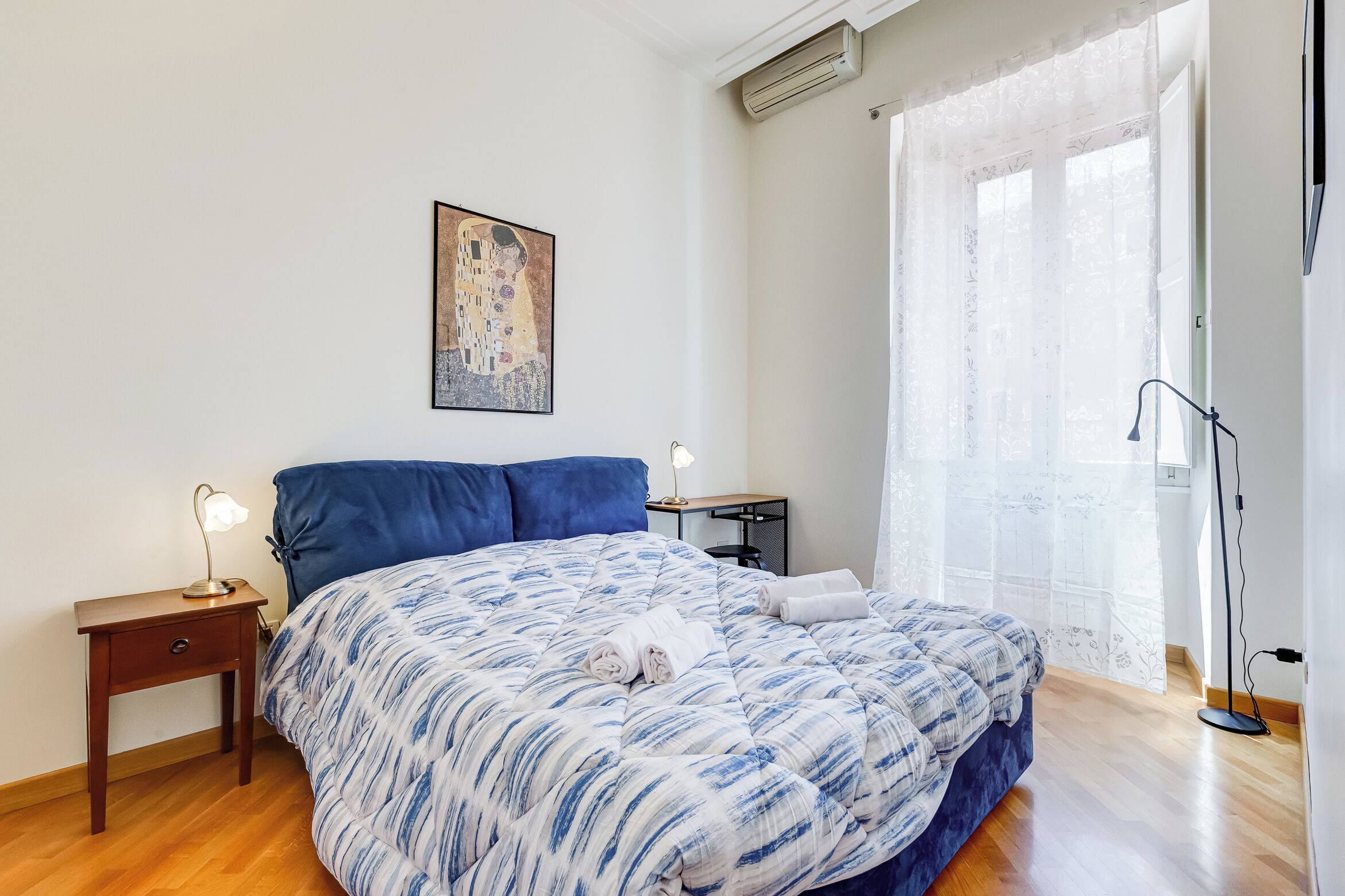 Een slaapkamer met een groot bed, een nachtkastje met een lamp, een kunstwerk in een lijst aan de muur en een raam met dunne gordijnen.