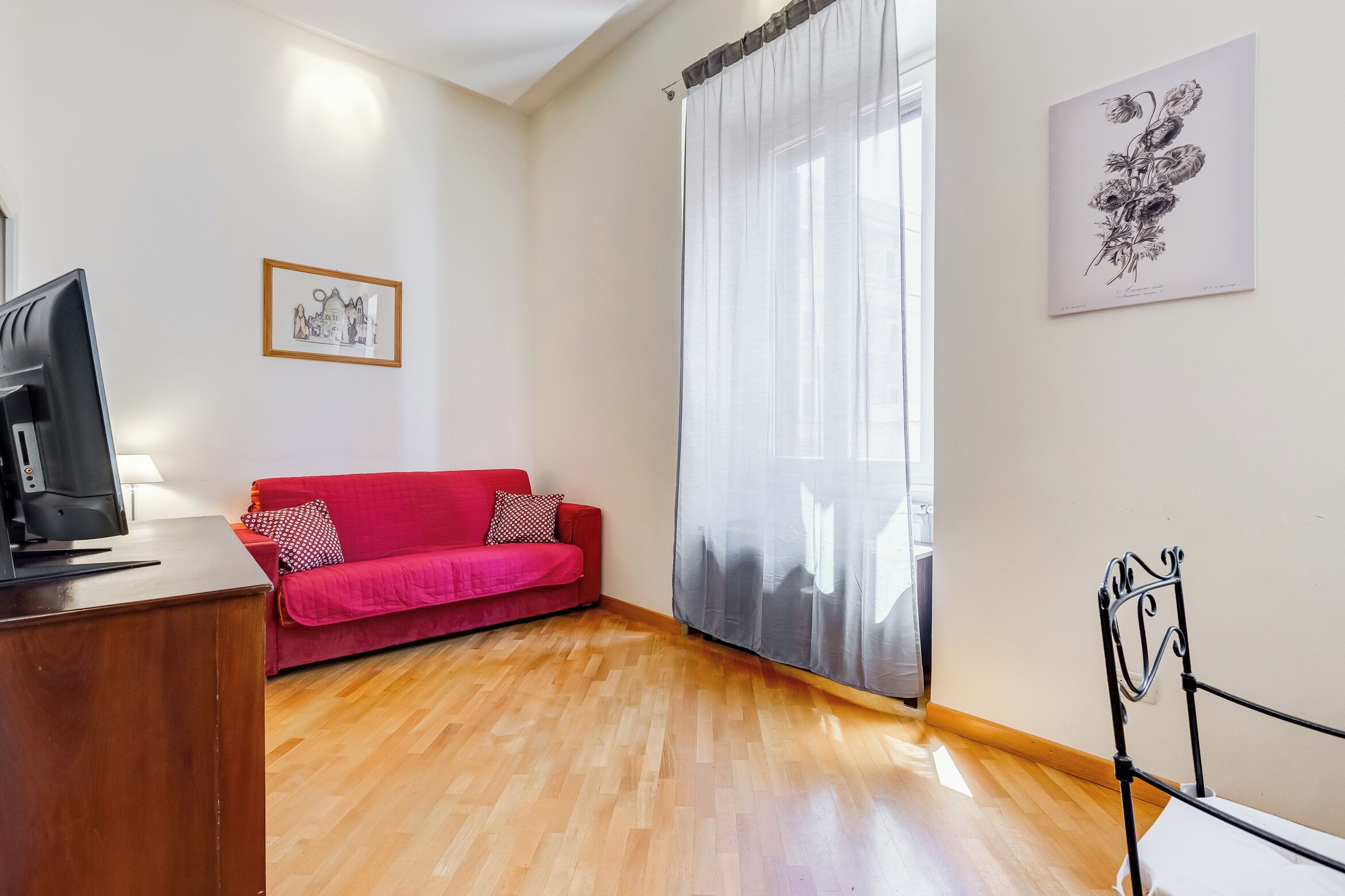 Une pièce avec un plancher en bois, un canapé rouge, un téléviseur posé sur un meuble en bois, un lit avec un cadre en métal et une fenêtre ornée de rideaux légers.