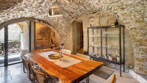 Ein rustikaler Innenraum aus Stein mit einem Holzespeisetisch, Stühlen, einem Vitrinenschrank mit Geschirr und einer Hängelampe.