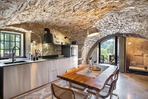 Une cuisine rustique aux murs de pierre, avec une table à manger en bois et une vue sur l’extérieur, visible à travers une porte en arc de cercle.