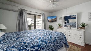 Un dormitorio con una cama grande, ventilador de techo, ventana con cortinas, cómoda y televisor instalado en la pared.