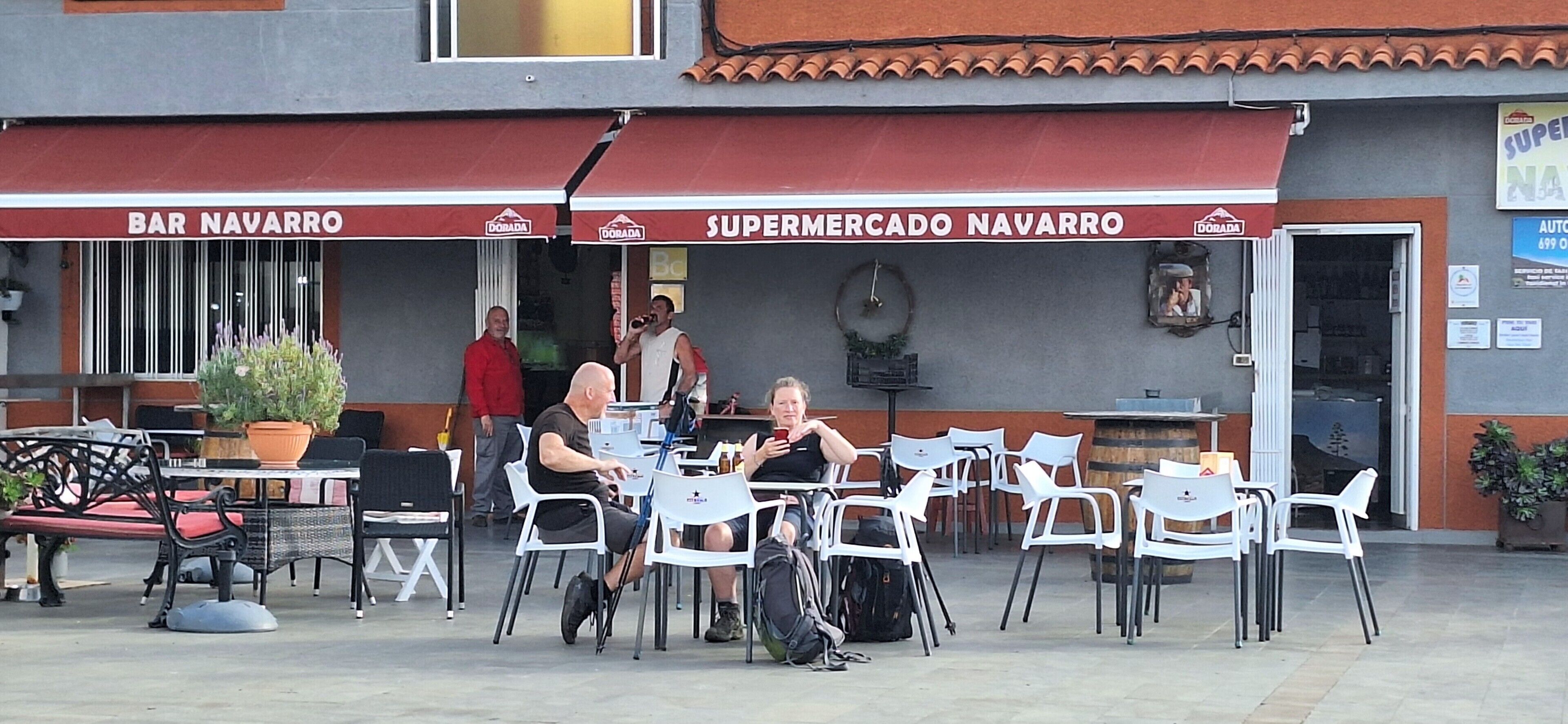 Un espace extérieur aménagé avec des tables et des chaises, un pot de fleurs et un bâtiment sur lequel on peut lire les enseignes « BAR NAVARRO » et « SUPERMERCADO NAVARRO ».
