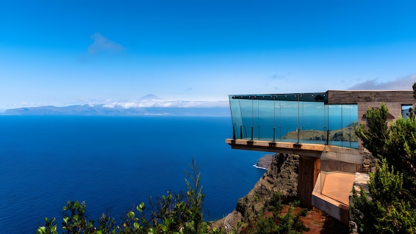 Eine Glasbrücke erstreckt sich über eine Klippe mit Blick auf das Meer und Berge im Hintergrund.