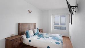 Une chambre à coucher avec un lit, des tables de chevet en bois, un téléviseur fixé au mur et une fenêtre donnant sur un beau paysage.