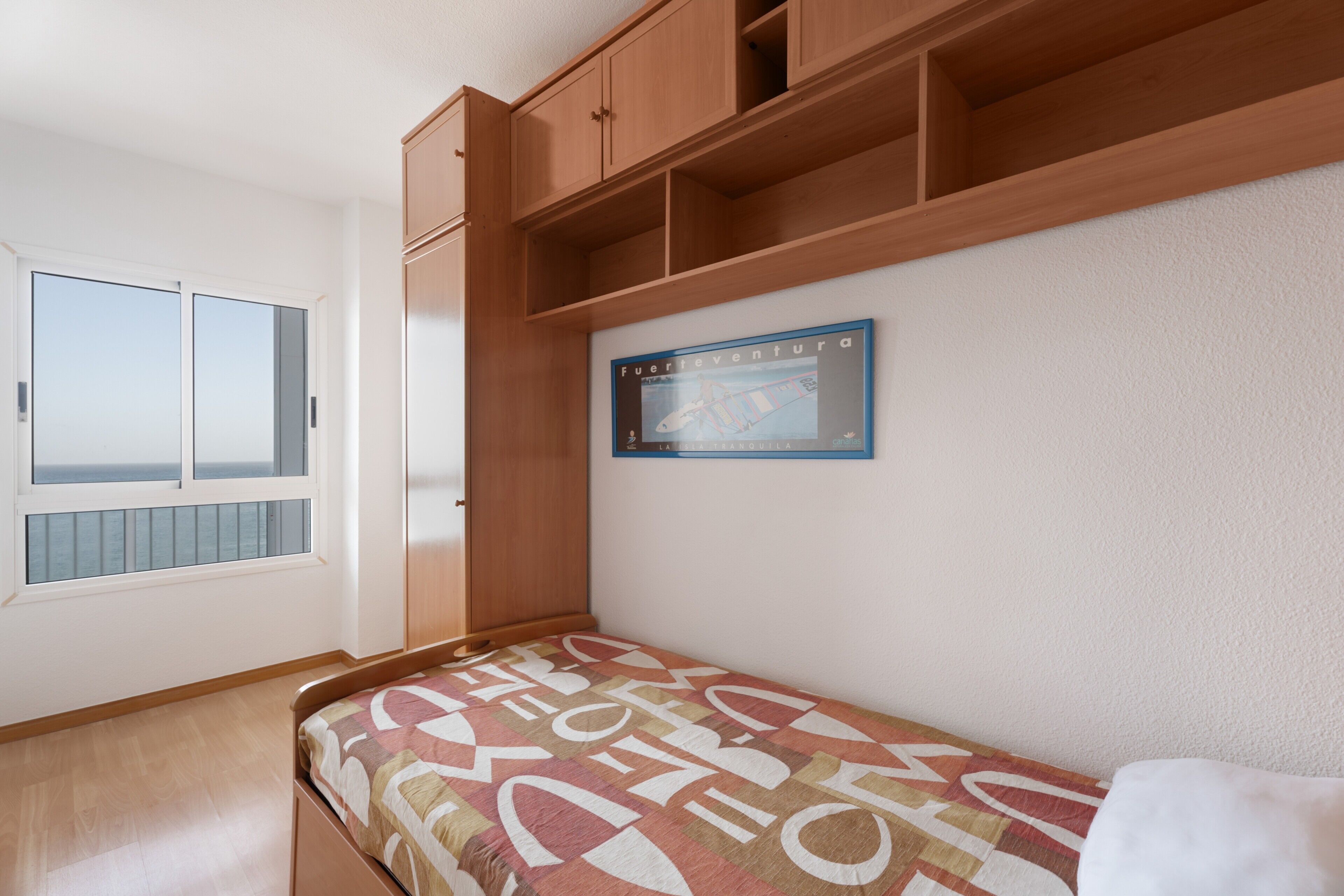 A bedroom with a bed, wooden cabinets, a window with a view, and a framed picture on the wall.