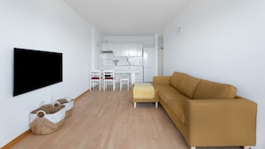 Un salon moderne avec un téléviseur à écran plat, un canapé, une table à manger avec des chaises, et un coin cuisine à l’arrière-plan.