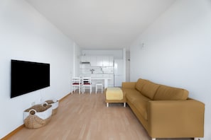 A modern living room with a flat-screen TV, a sofa, a dining table with chairs, and a kitchen area in the background.
