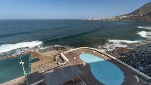 Une zone de baignade en bord de mer, dotée de deux piscines, d’une terrasse en bois et offrant une vue sur l’océan et les bâtiments situés au loin.