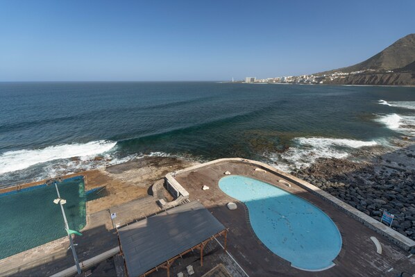 A coastal swimming area with two pools, a wooden deck, and a view of the ocean and distant buildings.