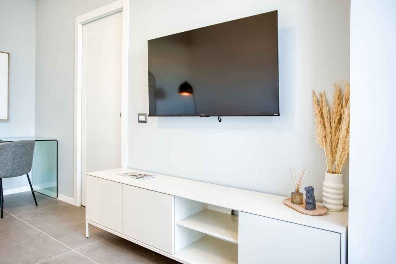 Un soggiorno moderno con una TV a schermo piatto montata a parete, un tavolino consolle bianco con oggetti decorativi e una poltrona in vetro.