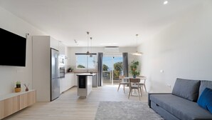 Ein modernes Wohnzimmer mit grauem Sofa, einem Flachbildfernseher an der Wand, einer Küchenzeile mit Kühlschrank und einem Essbereich mit Tisch und Stühlen.