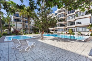 Un área de piscina con sillones reclinables, un árbol y un edificio con balcones.