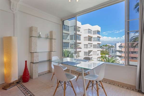 Un espace repas moderne avec une table en verre, des chaises blanches et une grande fenêtre donnant sur des immeubles résidentiels.