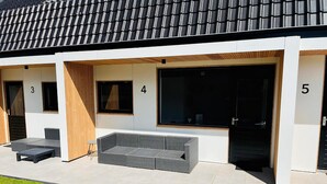 Edifici modern amb habitacions numerades 3, 4 i 5. Cada habitació té un gran finestral i una porta. A l'exterior hi ha una zona de seients amb una banca i una taula petita.