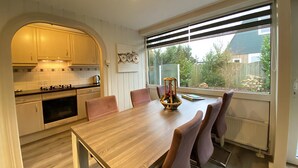 A modern kitchen with white cabinets, a wooden dining table, and a view of the backyard through large windows. - Verhuurburo Callantsoog - De Garnekuul 4 (Callantsoog)