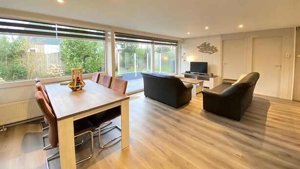 A modern living room with a wooden dining table, black sofas, and a TV. Large windows offer a view of the garden. - Verhuurburo Callantsoog - De Garnekuul 4 (Callantsoog)