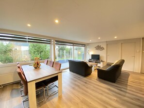 Un salon moderne avec une table à manger en bois, des canapés noirs et un téléviseur. De grandes fenêtres offrent une vue sur le jardin.