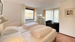 Ein ordentlich gemachtes Bett mit Kissen, eine Nachttischlampe, ein Schminktisch, ein Stuhl und ein Fenster mit Vorhängen.