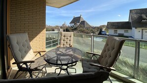 A balcony with a round table and four chairs, a glass railing, and a view of a house and trees. - Callantsoger Staete A107 (Callantsoog)