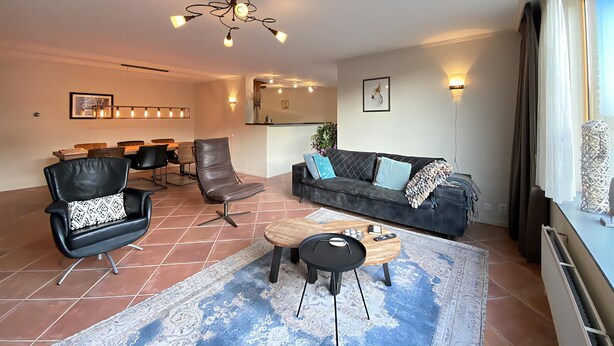 A modern living room with a black leather sofa, a wooden coffee table, and a patterned rug. There is a dining area in the background with a table and chairs. - Callantsoger Staete A107 (Callantsoog)