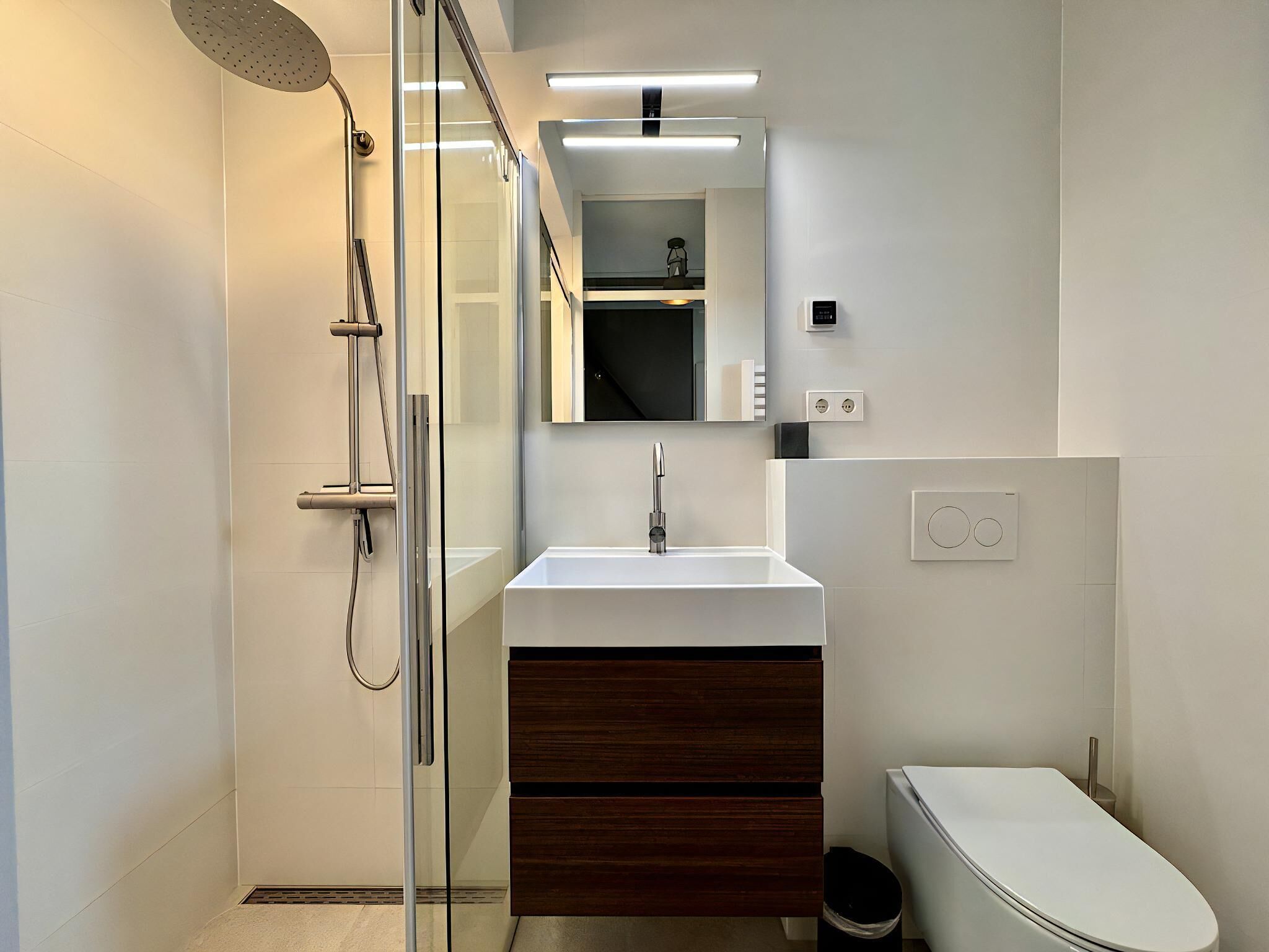 Ein modernes Badezimmer mit einer gläsernen Duschkabine, einem weißen Waschbecken mit Holzunterschrank und einer Toilette.