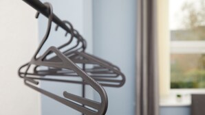 Un tendedero metálico con múltiples perchas, fijado a una pared en una habitación con una ventana y una planta a la vista.