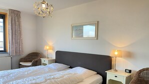 Un lit bien fait, avec du linge de lit blanc, deux tables de chevet avec des lampes, une chaise en osier et un tableau encadré au mur.