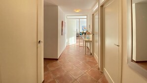 Corredor com portas brancas e chão de azulejos, que leva a um quarto com escrivaninha e luminária.