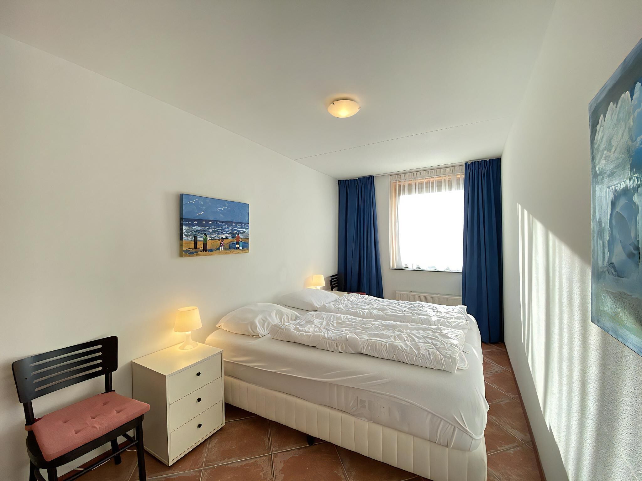 A bedroom with a bed, a nightstand, a chair, a painting, and a window with curtains.