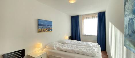 A bedroom with a bed, a nightstand, a chair, a painting, and a window with curtains.