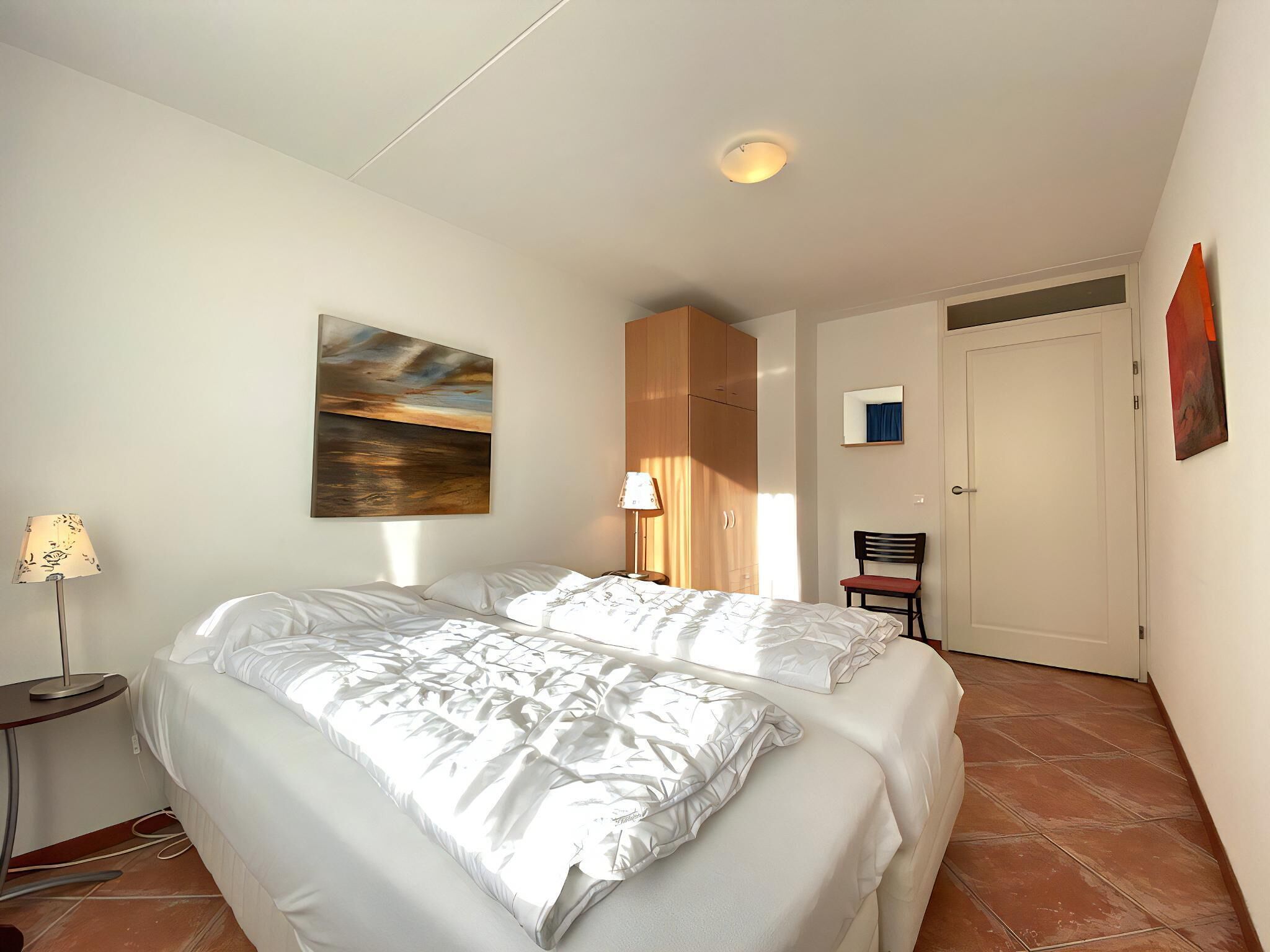Un dormitorio con cama, mesita de noche, lámpara, silla y puerta.
