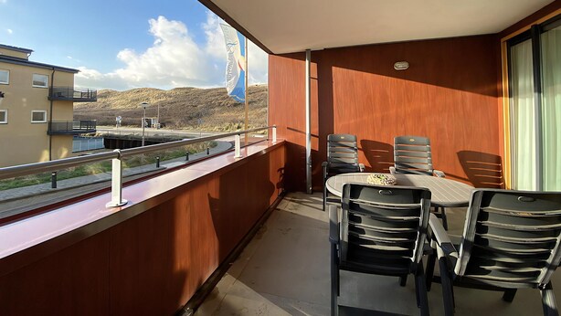 Uma varanda com mesa e cadeiras, vista para um edifício e montanhas, e uma bandeira.