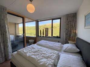 A bedroom with a large bed, a window with a view of a green landscape, and a balcony with a chair and table.