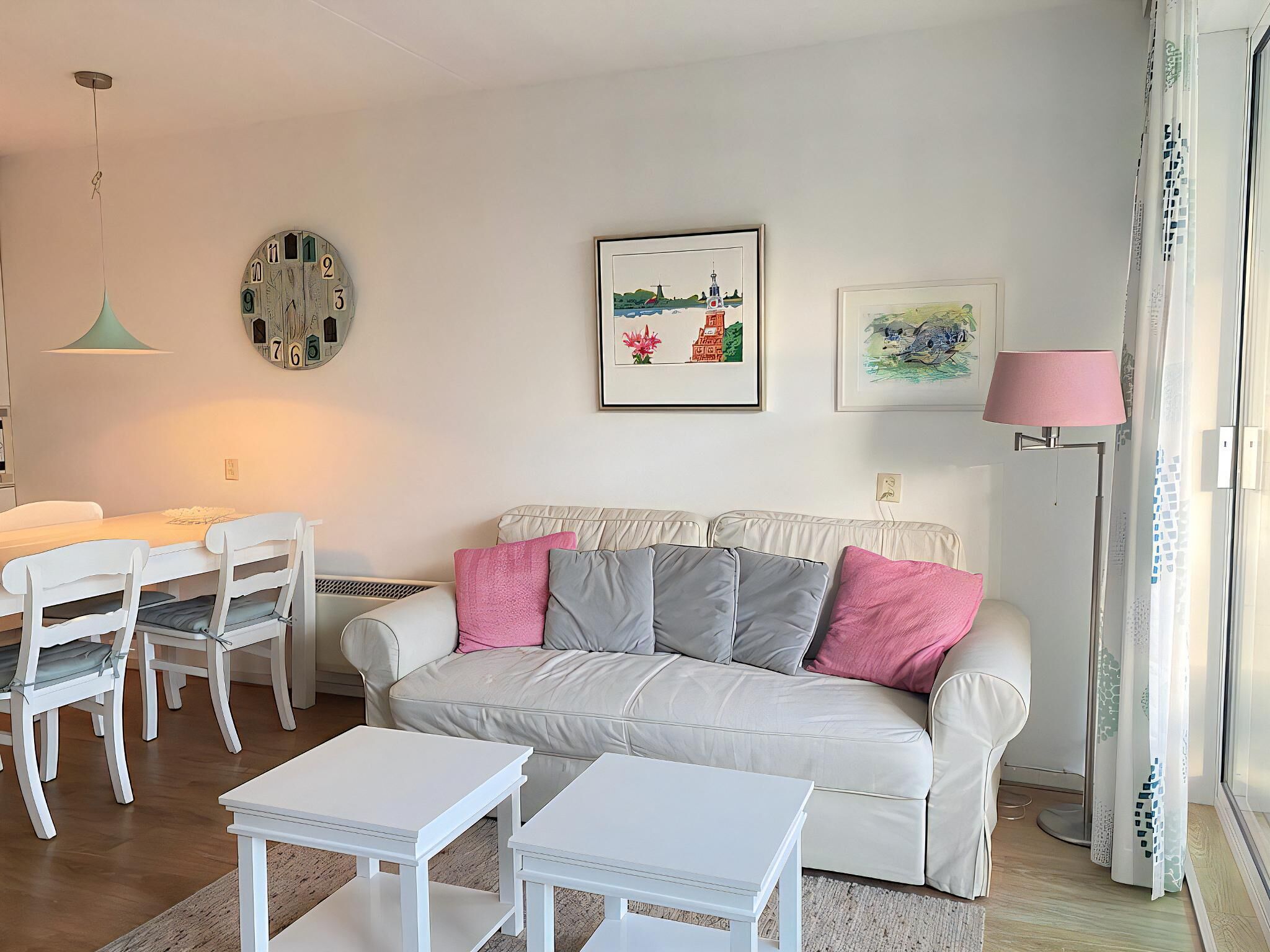 A living room with a white sofa, two white side tables, a dining table with chairs, a clock, and framed artwork on the walls.