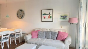 A living room with a white sofa, two white side tables, a dining table with chairs, a clock, and framed artwork on the walls.