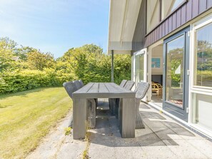 An outdoor patio with a wooden table and wicker chairs, surrounded by a well-maintained lawn and dense greenery. - 3 bedroom accommodation in Callantsoog (Callantsoog)