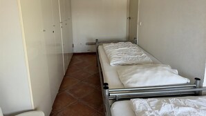 Un couloir étroit, avec un lit sur la droite, une armoire sur la gauche et un sol carrelé.