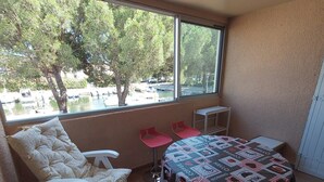 A small room with a bed, two chairs, a table, and a window with a view of boats and trees.